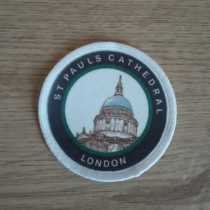 May include: A white, round patch with black and green trim. The patch features a drawing of St Paul's Cathedral in London. The text "St Paul's Cathedral London" is printed on the patch.