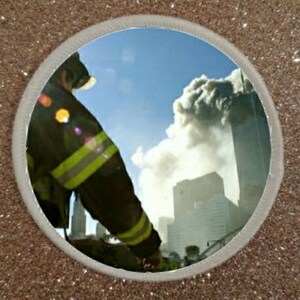 May include: A firefighter in a dark uniform with yellow reflective stripes stands in front of a large cloud of smoke and a tall building. The building is partially obscured by the smoke.