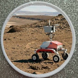 May include: A red and white rover with a NASA logo on the side is driving on a rocky, brown surface. The rover has six wheels and a large antenna on top.