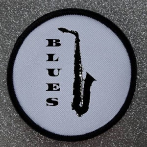 Pode incluir: Etiqueta redonda branca com borda preta apresentando uma silhueta preta de um saxofone e a palavra "BLUES" em letras pretas.