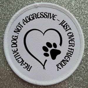 Puede incluir: Parche redondo blanco con texto negro y una huella de pata dentro de un corazón. El texto dice "Perro reactivo, no agresivo - simplemente demasiado amigable".