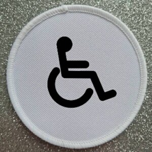 May include: A round, white patch with a black wheelchair symbol. The symbol is centered on the patch, which has a white stitched border. The background is a sparkly, silver surface.