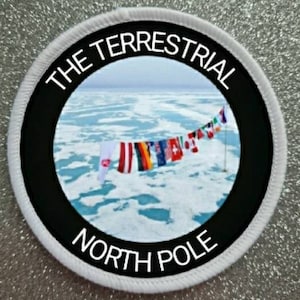 Könnte beinhalten: Ein weißer runder Patch mit schwarzem Text und schwarzem Rand. Der Text lautet "The Terrestrial North Pole". Innerhalb des Randes befindet sich ein Foto eines zugefrorenen Ozeans mit einer Reihe von Flaggen, die an einer Leine hängen.