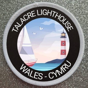 Może przedstawiać: Okrągła naszywka w kolorze białym i czarnym z napisem "Talacre Lighthouse Wales - Cymru" wokół krawędzi. Środek naszywki przedstawia kolorową ilustrację latarni morskiej i żaglówki na wodzie.