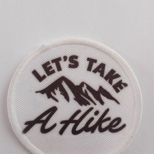 May include: White round patch with brown stitching around the edge. The patch has a brown mountain range graphic and the text "Let's Take A Hike" in a brown cursive font.