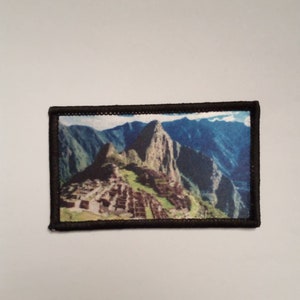 May include: A brown and black embroidered patch with a colorful image of Machu Picchu, an ancient Incan city in Peru.