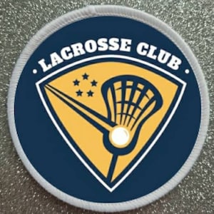 Puede incluir: Un parche circular blanco con fondo azul y un diseño amarillo de palo de lacrosse y pelota. El texto "LACROSSE CLUB" está escrito en blanco alrededor del borde del parche.