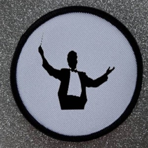 Könnte beinhalten: Ein weißer runder Patch mit schwarzem Rand, der die Silhouette eines Dirigenten mit einem Taktstock in der Hand und ausgestreckten Armen zeigt.