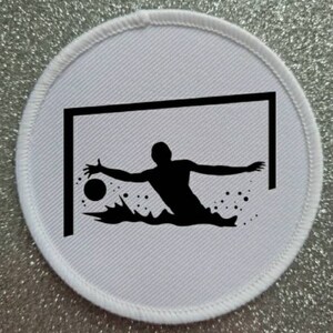 May include: A white round patch with a black silhouette of a soccer player diving to save a ball in a goal. The player is in the water and the ball is in the air.