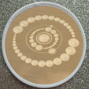 Könnte beinhalten: Ein beiger runder Patch mit weißem Rand. Der Patch zeigt ein weißes Spiraldesign mit konzentrischen Kreisen.
