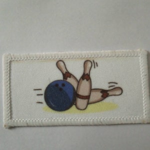 May include: White fabric patch with a blue bowling ball and brown bowling pins. The bowling ball is rolling towards the pins, which are knocked down.