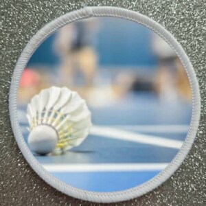 May include: A white badminton shuttlecock lying on a blue badminton court with white lines. The shuttlecock is in focus, while the court and the background are blurred.