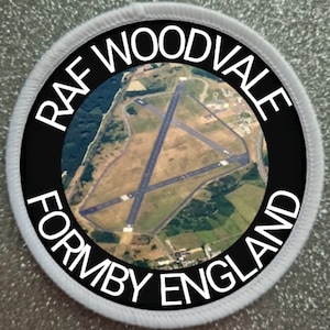 Può includere: Un cerotto circolare bianco e nero con il testo "RAF WOODVALE FORMBY ENGLAND" attorno al bordo. Il centro del cerotto presenta una fotografia aerea di una pista di atterraggio.