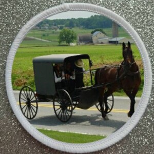 Könnte beinhalten: Ein schwarzer, von einem Pferd gezogener Wagen mit einer grauen Holzkiste und schwarzen Rädern. Der Wagen wird von zwei Personen gefahren, die traditionelle Kleidung und Hüte tragen. Der Wagen fährt auf einer asphaltierten Straße mit grünem Gras und Bäumen auf beiden Seiten.