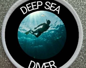 Könnte beinhalten: Ein weißer runder Patch mit einem schwarzen Kreis in der Mitte. Der schwarze Kreis trägt den Text "DEEP SEA DIVER" in weißen Buchstaben. Im Inneren des schwarzen Kreises befindet sich ein Foto eines Tauchers, der unter Wasser schwimmt.