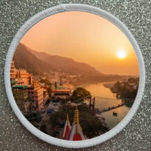 Puede incluir: Un parche redondo blanco con una imagen panorámica de una ciudad enclavada en un valle al atardecer. La ciudad está rodeada de montañas y un río fluye por el valle. El sol se pone detrás de las montañas, proyectando un cálido resplandor sobre la escena.