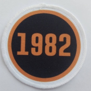 May include: A white circular patch with an orange border and a black circle in the center. The black circle has the number 1982 printed in orange.