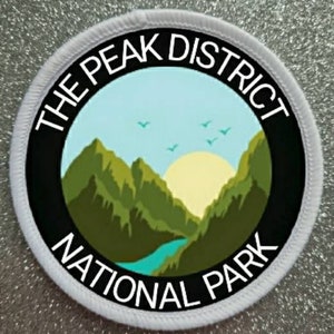 Op de afbeelding: Ronde witte patch met zwarte tekst en een groene en blauwe berg scène. De tekst luidt "The Peak District National Park".
