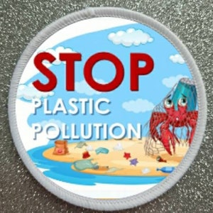 Op de afbeelding: Een ronde witte patch met een blauwe achtergrond en een rode banner met de tekst "STOP PLASTIC POLLUTION". Een cartoonkrab is gevangen in een net met plastic afval op een strand.
