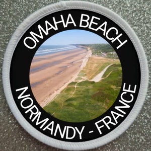 May include: A black and white circular patch with the text "Omaha Beach Normandy - France" around the edge. The center of the patch features a photograph of a beach with sand dunes and green grass.