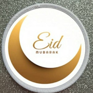 Puede incluir: Parche redondo blanco con una luna creciente dorada y el texto "Eid Mubarak" en letras doradas.