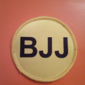 Puede incluir: Un parche circular amarillo con costuras negras alrededor del borde. El parche tiene las letras "BJJ" impresas en negro.