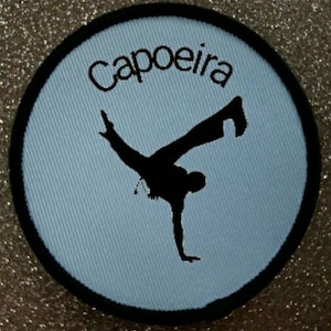 Könnte beinhalten: Ein runder blauer und schwarzer Patch mit dem Wort "Capoeira" in schwarzer Stickerei. In der Mitte des Patches befindet sich eine schwarze Silhouette einer Person, die eine Capoeira-Bewegung ausführt.