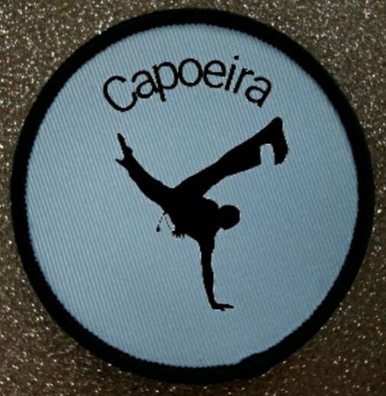 K&ouml;nnte beinhalten: Ein runder blauer und schwarzer Patch mit dem Wort "Capoeira" in schwarzer Stickerei. In der Mitte des Patches befindet sich eine schwarze Silhouette einer Person, die eine Capoeira-Bewegung ausf&uuml;hrt.