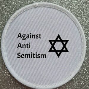 Puede incluir: Parche redondo blanco con texto negro que dice "Against Anti Semitism" y una estrella de David negra en el centro.