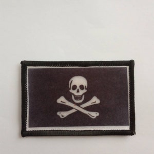 May include: A rectangular patch featuring a white skull and crossbones on a black background. The patch has a black border and is likely an emblem or insignia, possibly for a pirate-themed item.
