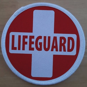May include: A white and red round patch with a white cross and the word "LIFEGUARD" in red letters.