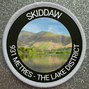 May include: White round patch with black border and a photo of a mountain range and lake. The text "SKIDDAW 931 METRES - THE LAKE DISTRICT" is printed in white around the photo.