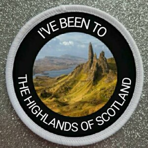 Op de afbeelding: Een ronde witte en zwarte patch met de tekst "I've Been To The Highlands Of Scotland" en een foto van een rotsachtig berggebied met een blauw meer op de achtergrond.