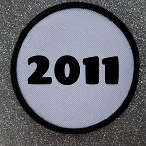Könnte beinhalten: Ein runder Aufnäher mit hellblauem Zentrum und schwarzem Rand. Die Zahl "2011" ist in fetter schwarzer Schrift über die Mitte des Aufnähers gedruckt. Der Hintergrund ist silbern glitzernd.