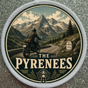 Puede incluir: Un parche bordado circular que muestra a un motociclista recorriendo los Pirineos. La escena incluye una carretera sinuosa, un hito de piedra con la inscripción COL D'AUBISQUE 1709m y el texto THE PYRENEES. El diseño tiene un borde blanco.