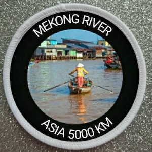 May include: Circular patch with the text "MEKONG RIVER" and "ASIA 5000 KM". The patch features a scenic image of a person rowing a boat on a river, with houses on the riverbank. The patch has a black border with white text.