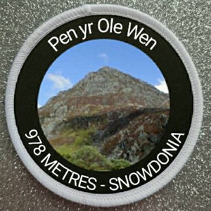 Può includere: Un cerotto rotondo bianco con testo nero e una foto di una montagna. Il testo recita "Pen yr Ole Wen 978 Metres - Snowdonia".