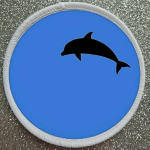 May include: A white round patch with a blue background and a black silhouette of a dolphin jumping out of the water.