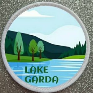 Könnte beinhalten: Ein runder weißer Patch mit einer blauen und grünen Illustration eines Sees und Bergen. Der Text "LAKE GARDA" ist in grünen Buchstaben auf dem Patch geschrieben.