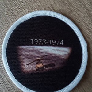 Puede incluir: Un parche circular blanco con fondo negro y borde blanco. El parche presenta un helicóptero volando sobre una superficie lunar. El texto "1973-1974" está impreso en blanco en el parche.