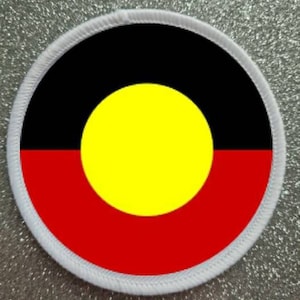 May include: A white circular patch with a red, yellow and black Aboriginal flag design. The flag features a yellow circle in the centre, surrounded by a black ring, and a red band at the bottom.