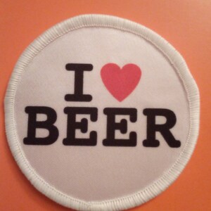 Könnte beinhalten: Runder weißer Patch mit schwarzem Rand und dem Text "I love BEER" mit einem roten Herzen zwischen den Wörtern.