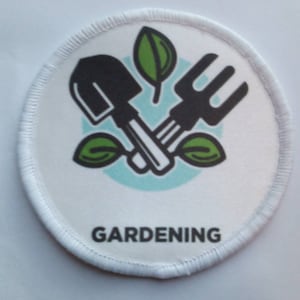 Könnte beinhalten: Runder weißer Aufnäher mit einem grünen Blattdesign und einer schwarzen Schaufel und Harke. Der Text "GARDENING" ist unter dem Design gedruckt.