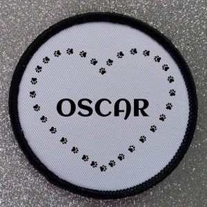Può includere: Cerotto rotondo bianco con bordo nero. Il cerotto ha una forma di cuore fatta di impronte di zampe con il nome "OSCAR" all'interno.
