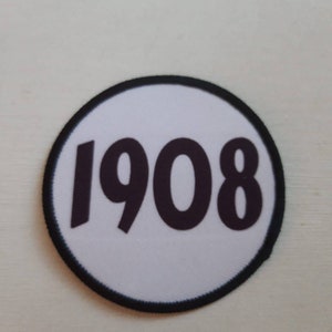 May include: A white circle patch with a black border and the number 1908 printed in black.