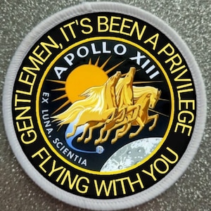 Pode incluir: Um remendo redondo preto e amarelo com o texto "Gentlemen, it's been a privilege flying with you." O remendo também apresenta o texto "Apollo XII" e um gráfico de três cavalos dourados correndo sobre a lua.