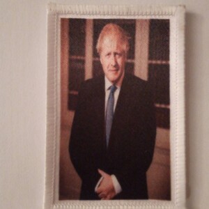 Peut inclure: Un écusson en tissu blanc avec une bordure cousue représentant une photographie couleur d'un homme en costume et cravate. L'homme a les cheveux blonds et sourit.
