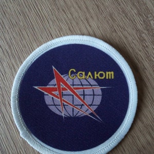 May include: A round, blue patch with a white border. The patch features a red star with a white globe inside. The text "Салом" is written in yellow letters above the star.