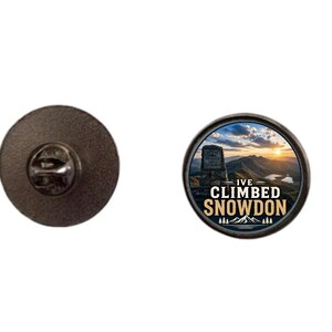 May include: A round pin with a dark metal finish. The pin features a scenic image of a mountain range with the text "I'VE CLIMBED SNOWDON". The pin has a pin back for attachment.