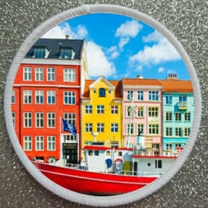 Può includere: Una barca rossa con una cabina bianca è ormeggiata davanti a una fila di edifici colorati. Gli edifici sono dipinti in tonalità di rosso, giallo, rosa e blu. Il cielo è blu con nuvole bianche.